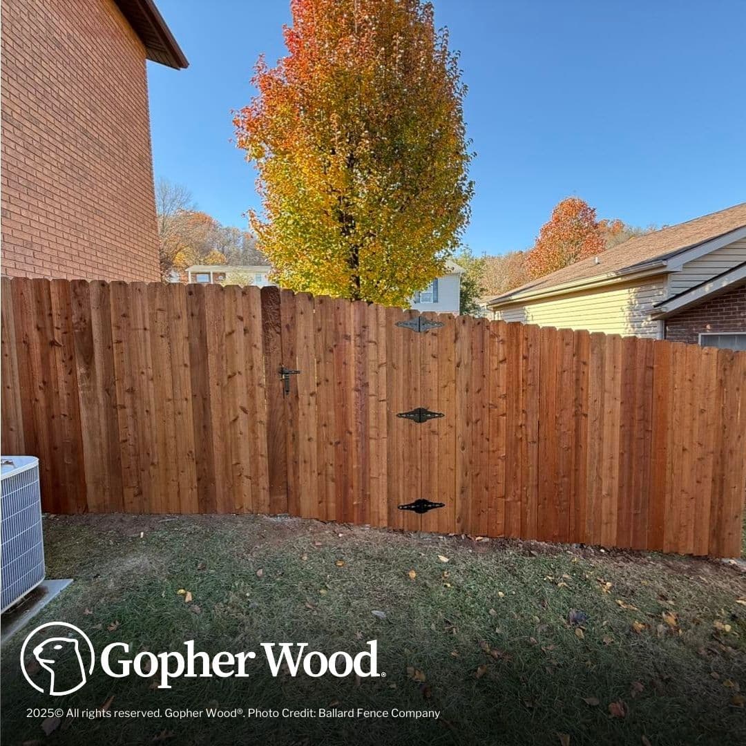 Wooden privacy fence and gate with black hinges against a colorful autumn tree backdrop.
