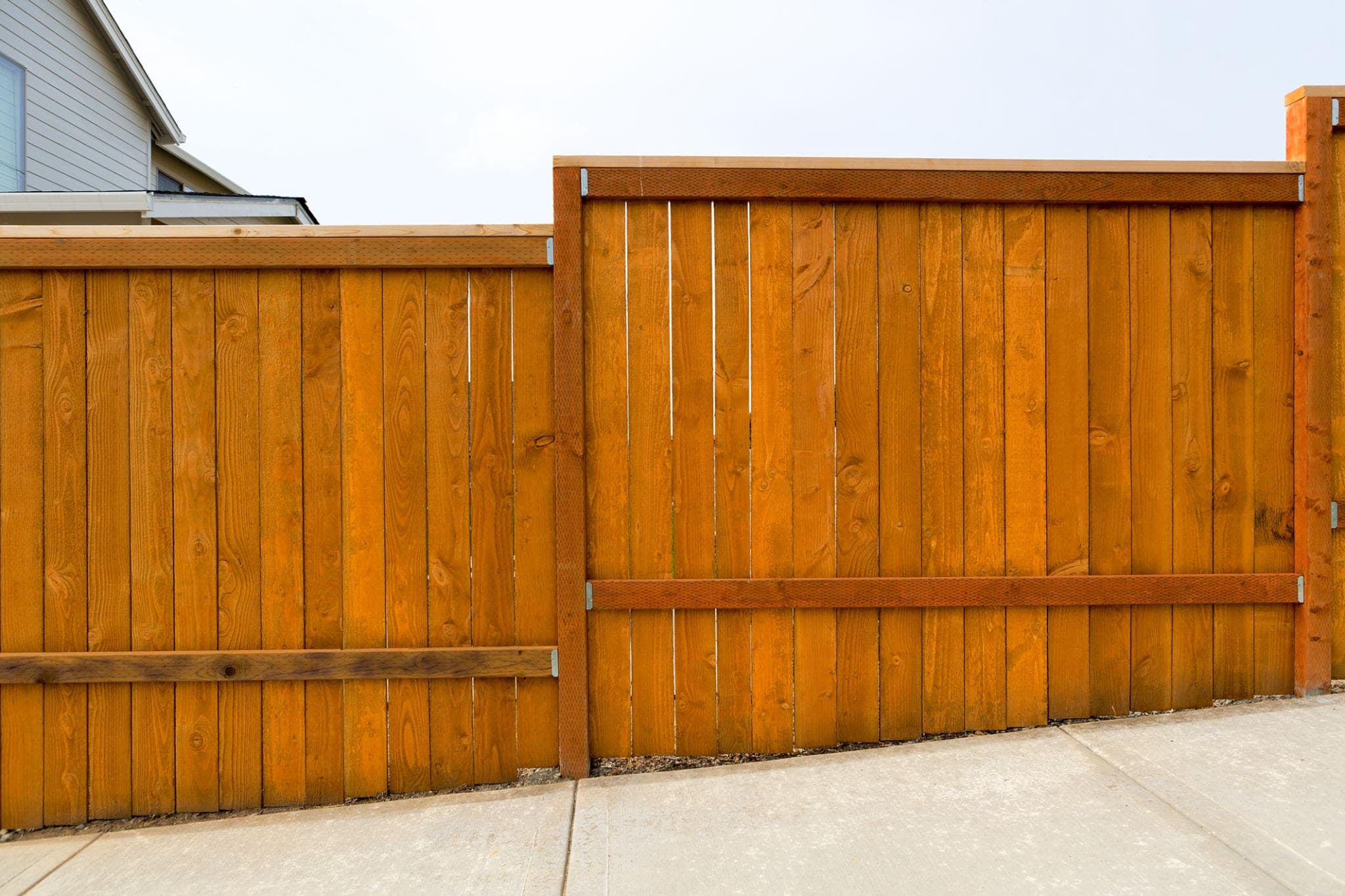 Orange-brown stained wooden fence with vertical slats bordering a concrete sidewalk on a slope.